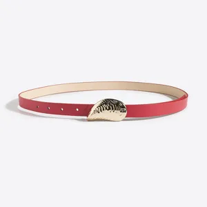 Detalle de cinturón fino de cuero rojo para mujer con hebilla dorada en forma de concha marina.