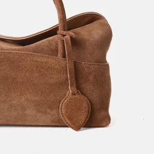Gros plan sur la poignée et le charm du cabas en suédine marron, texture de cuir premium.