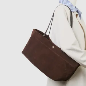 Detalle de bolso tote de ante marrón, textura suave, correas de hombro finas y botón metálico.
