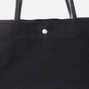 Detalle de bolso tote de lona negra con textura de tela, asas de cuero y botón dorado.