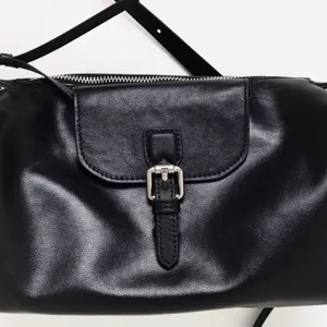 Vista detallada de bolso de cuero negro para mujer bajo el brazo con hebilla y cierre de cremallera.