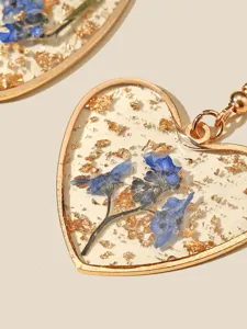 Nahaufnahme von herzförmigen Harzohrringen mit blauen Trockenblumen und dekorativer Goldfolie.