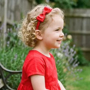 Close-up of a cute toddler girl wearing a vibrant red bow headband outdoors in garden setting.