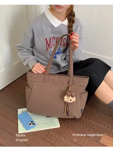 Sac brun à poches latérales et breloque ourson en peluche, tenu par un modèle assis dans une scène naturelle