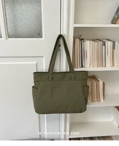 Cabas en nylon vert olive, texture plissée et poches latérales, suspendu près d’une bibliothèque