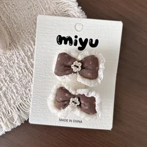 Dos pinzas de lazo marrón con encaje blanco y detalles florales color crema sobre una tarjeta