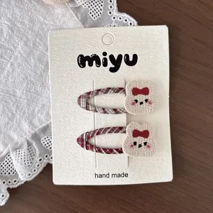 Close-up of two red plaid hair clips with cream knitted bear faces and red bows on a display card