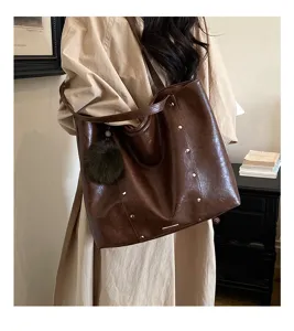 Model wearing a glossy brown tote with silver studs and a green pom-pom charm over one shoulder