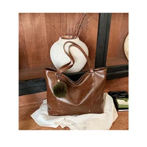 Brown glossy faux-leather tote with silver studs and a green pom-pom, styled on a white cloth