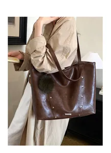 Model carrying a glossy brown tote with studded trim and a dark green pom-pom charm indoors