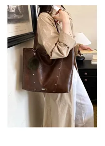 A model carries a brown bag over her shoulder, showing metal rivets and a fluffy charm indoors