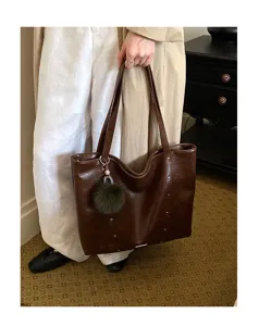 A model holds a glossy brown tote with double handles, small studs, and a green fuzzy bag charm