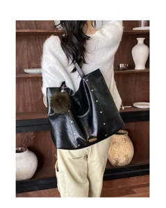 Black glossy faux-leather tote with metal studs and brown pom-pom charm, worn over the shoulder