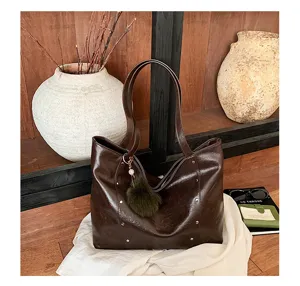 Brown faux-leather tote with metal studs and a green fuzzy charm, displayed against warm wood