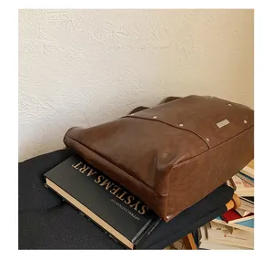 Angled view of a brown faux-leather tote with metal studs and a plaque, resting on books