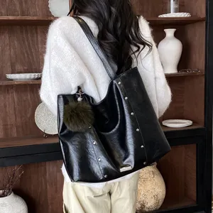 Black faux leather tote shoulder bag with stud accents and a pom-pom charm, worn from the back