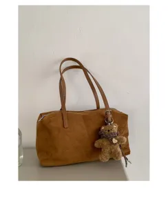 Bolso marrón de textura similar al ante con colgante de osito, ante una pared clara lisa