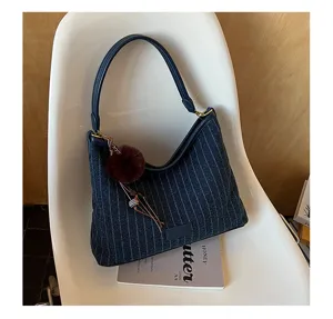 Gestreepte marineblauwe tas met leren hengsel, gouden details en bruine pompon op een witte stoel