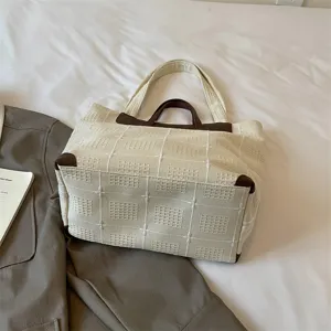 Cremefarbene Tasche mit Webstruktur und braunen Griffen auf weißer Bettwäsche neben grauem Mantel