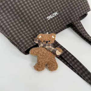 Close-up of a dark brown plaid tote bag with a plush teddy bear charm, checked scarf and heart tag