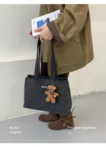 Lifestyle view of a dark gray checked tote with a teddy bear charm, carried beside a book
