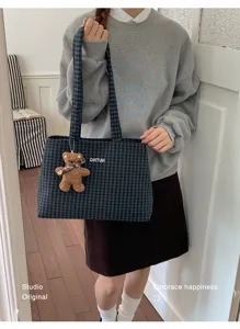 Person carries a blue-green plaid tote with a small bear charm, shown against an indoor wood floor.