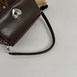 Close-up of a brown leather-look handbag with a silver heart clasp and curved strap on gray