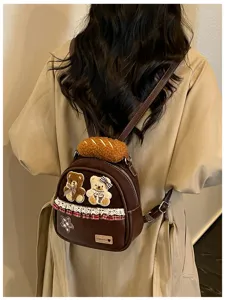 Back view showing a brown mini backpack with bear patches, plush bread charm, lace trim and star badge