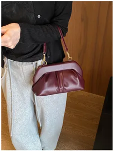 Borsa bordeaux arricciata con ganci dorati e tracolla sottile, portata davanti a uno sfondo di legno