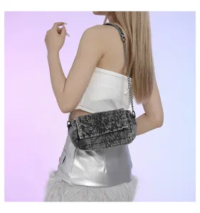 Modella con borsa a patta in denim grigio slavato e catena argentata, su uno sfondo sfumato delicato