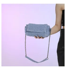 Dettaglio di una borsa blu in denim trapuntato, con patta e catena argentata, su sfondo lilla