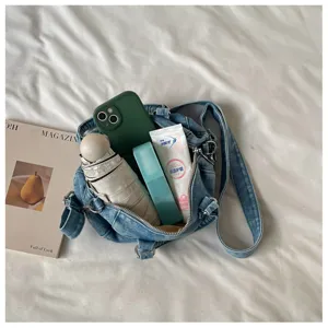 Vue à plat d’un sac en denim bleu délavé, avec bandoulière réglable, zip et objets à l’intérieur