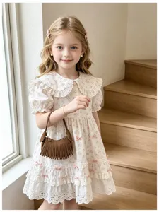 A child models a white floral dress while carrying a small brown fringed handbag indoors.