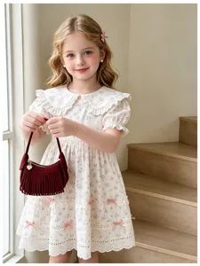 Child holding a burgundy mini handbag with long fringe tassels beside a white floral lace dress
