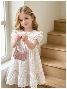 A child holds a blush-pink mini handbag with fringe beside a white floral dress near wooden stairs.