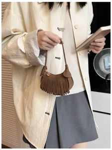 Close-up of a woman holding a brown suede-look fringed bag with a gold charm against a cream coat