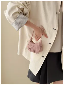 A hand holds a dusty pink suede mini bag with fringe and gold hardware against a neutral outfit