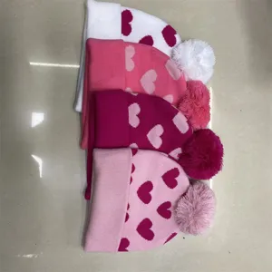 Four knitted beanies with heart jacquard patterns and fluffy pom-poms arranged on a light surface