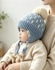 Baby wearing a blue knit hat with cream dots, fluffy pom-pom, earflaps and mitten-shaped ties
