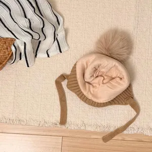Overhead view of a beige fleece-lined knit hat with a brown ribbed brim, pom-pom and ties