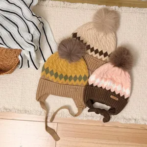 Three patterned knit hats with fluffy pom-poms, including tie-up earflaps, arranged on a cream rug