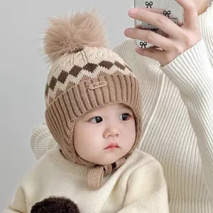 Baby wearing a beige and brown knit hat with pom-pom, diamond pattern, ear flaps and ties