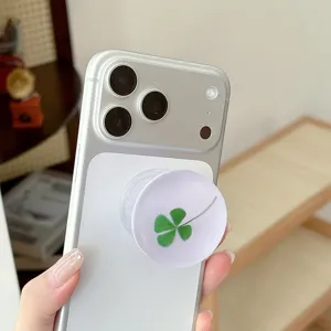 Een hand houdt een telefoon met witte ronde houder en groen klavertjevier vast, in huiselijke omgeving