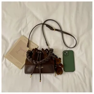 Flat lay showing a brown bucket bag with braided handle, drawstring closure, strap and bear charm