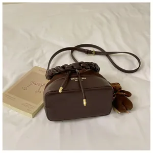 Brown bucket bag on white bedding, with braided handle, drawstring closure and bear charm
