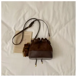 Flat lay detail of a dark brown suede bucket bag with leather trim, drawstring, hardware and bear charm