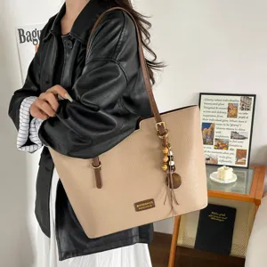 Cabas beige en similicuir grainé, bandoulière brun foncé et charm en bois, porté à l’épaule.