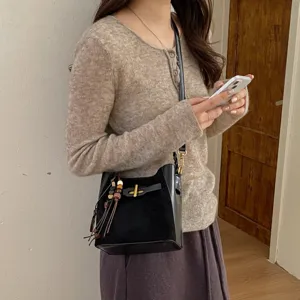 Black faux-leather bucket bag with gold clasp and wooden bead charm, worn crossbody by a model
