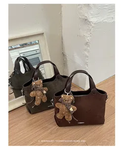 Three suede-textured tote bags in brown and olive, with plush bear charms, arranged on a wood floor