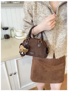 Sac brun à texture suédée, doté de deux anses et d’un pendentif en peluche, porté par une personne.